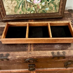 Puede incluir: Una bandeja de joyería de madera con tres compartimentos forrados con tela negra. La bandeja está hecha de madera de color claro y se encuentra sobre un baúl vintage. Una impresión floral enmarcada antigua está en el fondo.