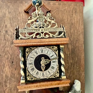 Op de afbeelding: Antieke wandklok met een donkere wijzerplaat, Romeinse cijfers en sierlijke goudkleurige wijzers. De klok heeft een houten frame met decoratieve metalen accenten en de tekst "NU-ELCK SYNESIN". Hij is op een houten oppervlak gemonteerd.