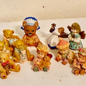 Puede incluir: Una colección de diez pequeñas figuritas de cerámica, incluyendo osos, ángeles y niños. Las figuritas están pintadas en varios colores, incluyendo azul, rosa, verde y marrón. Algunas de las figuritas sostienen pequeños objetos, como flores, estrellas y osos de peluche. Las figuritas están dispuestas sobre una superficie blanca.