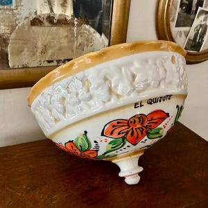 Puede incluir: Un cuenco de cerámica con base blanca, adornado con diseños florales naranjas y verdes. El borde es de color marrón claro. Las palabras "EL QUIJOTE" están escritas en el lateral. El cuenco tiene un pie decorativo.