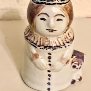 May include: A ceramic figurine of a woman in a white dress with a brown and blue hat. The figurine has a detailed face and a decorative collar. The dress is buttoned down the front, and a small figure is on the side.
