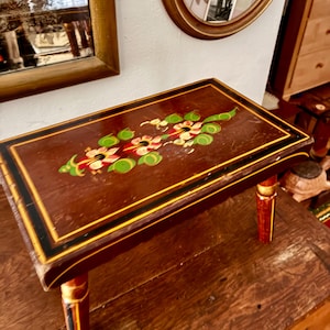 May include: A rectangular wooden footstool with a dark brown finish, featuring a hand-painted floral design in red, white, and green on the top surface. The stool has four cylindrical legs and is accented with black and yellow painted border lines.