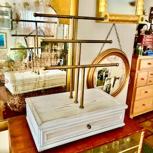 Peut inclure: Un organisateur de bijoux blanc avec une finition vieillie, comprenant un tiroir et un présentoir à barres en laiton à trois niveaux. L'organisateur repose sur une surface en bois, se reflétant dans un miroir. L'esthétique générale est vintage et élégante.