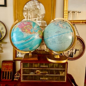 May include: Two vintage globes with blue oceans and colorful continents sit atop a wooden cabinet. The globes have gold-colored metal stands. A small wooden cabinet with multiple drawers is below the globes. The cabinet has the text "DMC" on it.