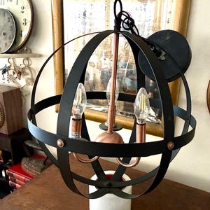 May include: Black metal orb chandelier with three light bulbs. The fixture has a copper-colored center and is mounted on a white base. The design features a spherical cage-like structure, suitable for home lighting.
