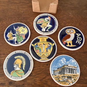 May include: Set of six ceramic coasters with Greek-themed designs, including an owl, a warrior, and the Parthenon. The coasters have a blue border and white trim. A matching beige coaster holder is also included.