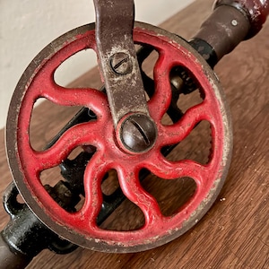 May include: Close-up of a vintage hand drill. The drill features a red spoked wheel, a dark metal handle, and a wooden grip. The metal components show signs of wear, suggesting age and use. The drill is set against a wooden surface.