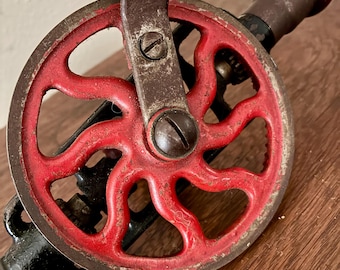 Puede incluir: Primer plano de un taladro manual antiguo. El taladro presenta una rueda de radios roja, un mango de metal oscuro y un agarre de madera. Los componentes metálicos muestran signos de desgaste, lo que sugiere edad y uso. El taladro está sobre una superficie de madera.