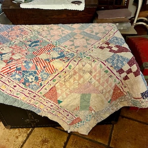 May include: An antique patchwork quilt with a variety of patterns and colors. The quilt features red, white, and blue fabrics with star and stripe designs, along with floral and geometric patterns. The quilt is draped over a dark trunk.