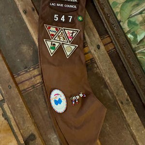 K&ouml;nnte beinhalten: Ein braunes Girl Scouts Halstuch mit Abzeichen und Anstecknadeln. Auf dem Halstuch steht "GIRL SCOUTS U.S.A. LAC BAIE COUNCIL" und die Nummer "547". Abzeichen zeigen verschiedene Designs, und auch Anstecknadeln sind vorhanden.