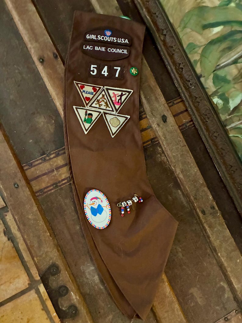 K&ouml;nnte beinhalten: Ein braunes Girl Scouts Halstuch mit Abzeichen und Anstecknadeln. Auf dem Halstuch steht "GIRL SCOUTS U.S.A. LAC BAIE COUNCIL" und die Nummer "547". Abzeichen zeigen verschiedene Designs, und auch Anstecknadeln sind vorhanden.