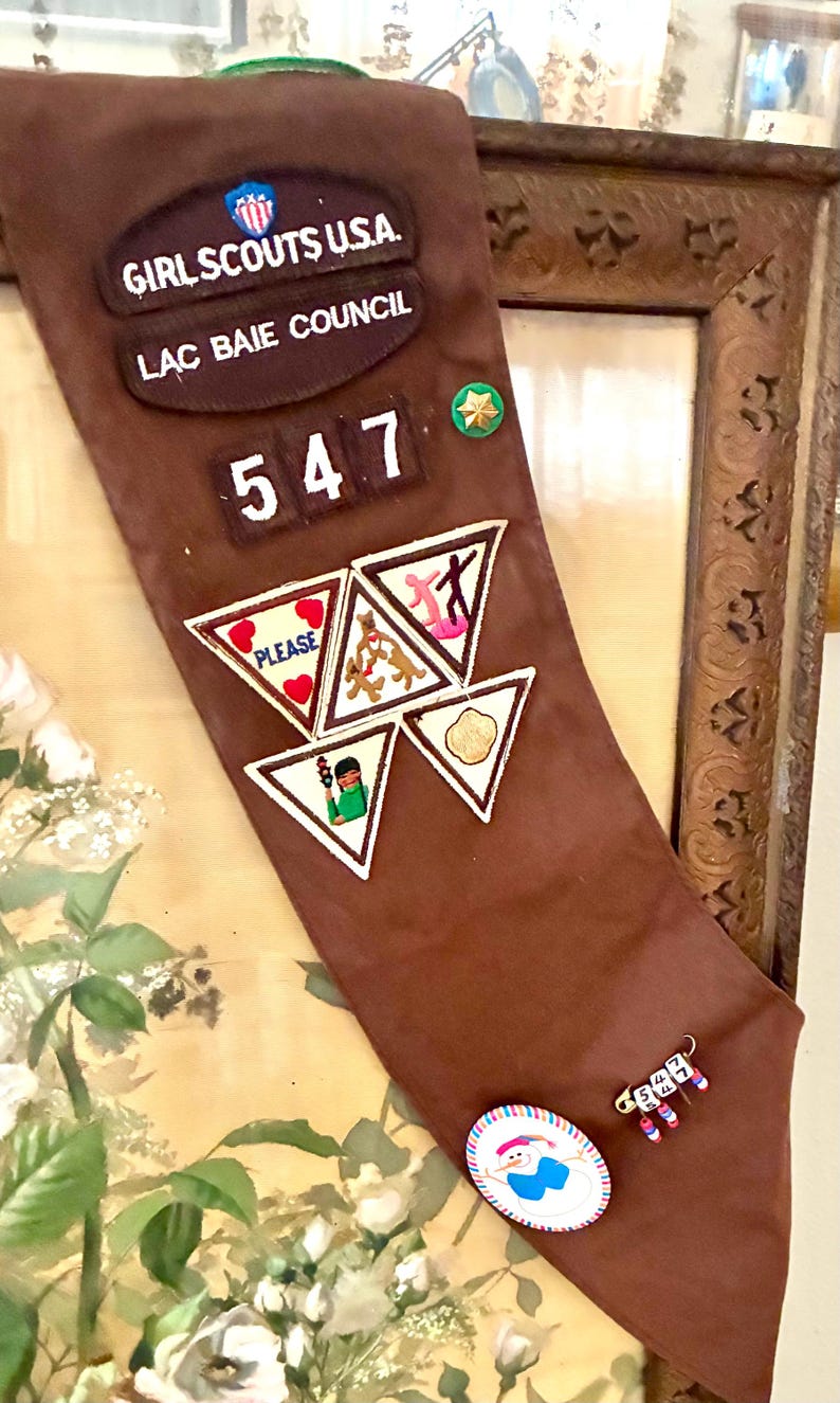 K&ouml;nnte beinhalten: Eine braune Girl Scouts Sch&auml;rpe mit Aufn&auml;hern und Anstecknadeln. Auf der Sch&auml;rpe steht "Girl Scouts U.S.A." und "Lac Baie Council". Zu den Aufn&auml;hern geh&ouml;ren ein Herz mit "Please" und Aktivit&auml;tssymbole. Eine Anstecknadel mit der Nummer "547" ist ebenfalls sichtbar.
