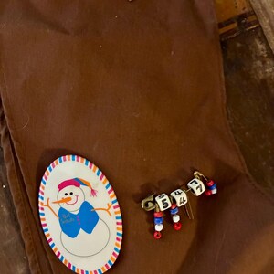 K&ouml;nnte beinhalten: Braune Weihnachtsstrumpf aus Stoff mit einer Schneemann-Anstecknadel und einer Anstecknadel mit Zahlenperlen. Die Schneemann-Anstecknadel hat einen blauen Schal und den Text "Girl Scouts". Die Zahlen-Anstecknadel hat die Zahlen 5, 4, 4 und 7.