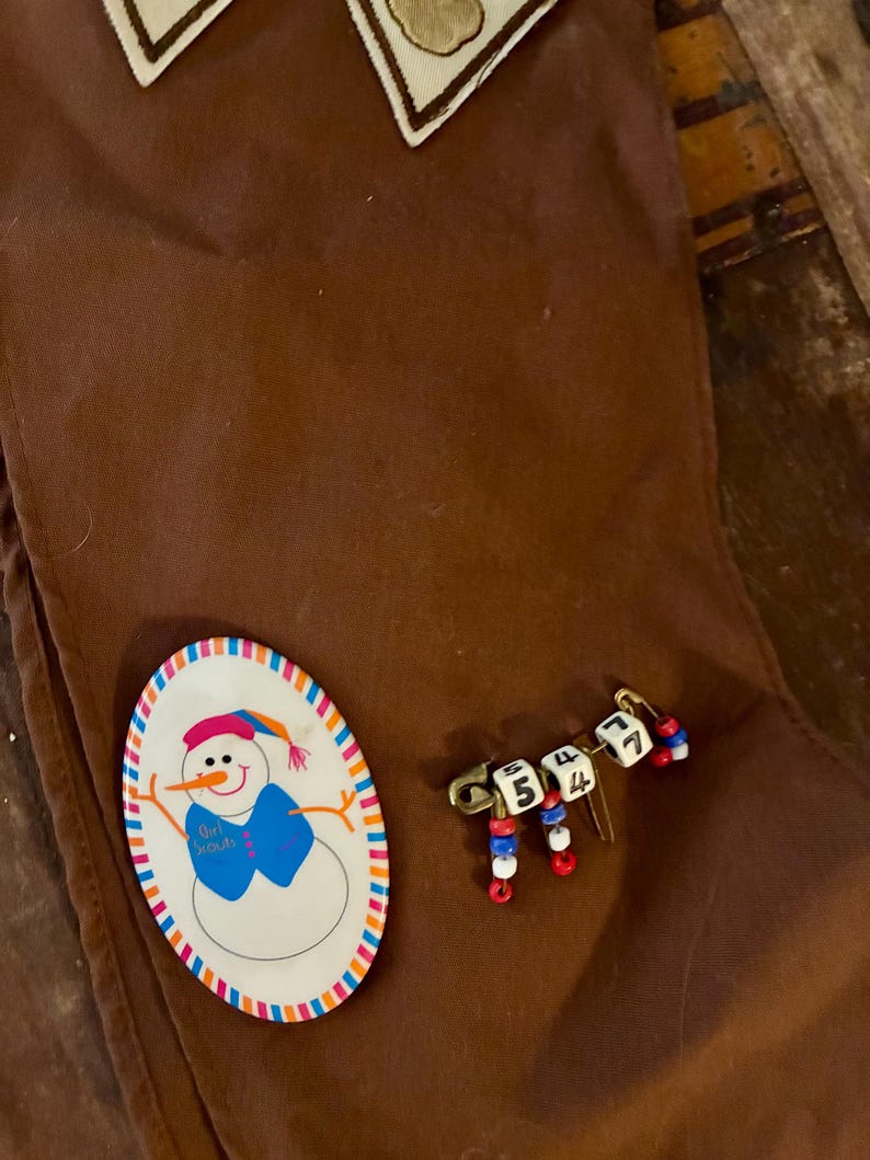 K&ouml;nnte beinhalten: Braune Weihnachtsstrumpf aus Stoff mit einer Schneemann-Anstecknadel und einer Anstecknadel mit Zahlenperlen. Die Schneemann-Anstecknadel hat einen blauen Schal und den Text "Girl Scouts". Die Zahlen-Anstecknadel hat die Zahlen 5, 4, 4 und 7.