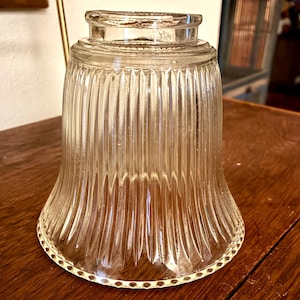 May include: Clear glass lampshade with vertical ribbed detailing. The lampshade has a flared shape and a decorative border around the bottom edge. The glass has a vintage appearance.