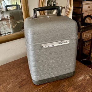 May include: A vintage grey Bell & Howell AutoLoad projector case with a textured finish, horizontal ribbed detailing, and a black plastic carrying handle on top. The front features a white rectangular label with the text Bell & Howell AutoLoad.