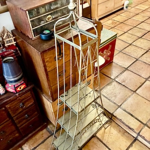 May include: A tall, off-white metal shelf unit with four shelves. The unit has a decorative, Eiffel Tower-inspired design. The shelves are rectangular and appear to be made of a light-colored material. The unit is placed on a tiled floor.