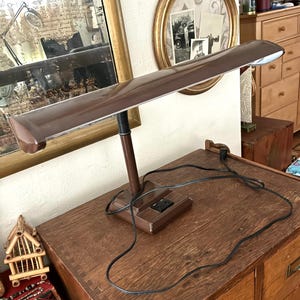 May include: A brown desk lamp with a rectangular shade and a flexible neck. The lamp has a square base with an on/off switch and a black power cord. The lamp is on a wooden surface.