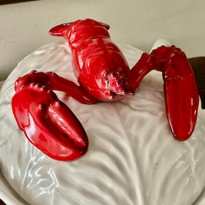 May include: A white ceramic tureen lid featuring a sculpted, glossy red lobster handle. The lid has a textured surface that mimics the appearance of overlapping cabbage leaves, with the lobster positioned centrally across the top of the lid.