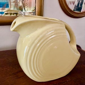 May include: A vintage, pale yellow pitcher with a curved handle and a spout. The pitcher has a rounded body with a series of concentric, ridged lines. The pitcher is sitting on a dark wooden surface.