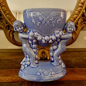 Puede incluir: Un jarrón de cerámica azul claro con un diseño detallado. Dos figuras de querubines sostienen una guirnalda de flores alrededor del centro del jarrón. El jarrón tiene una abertura ancha y se asienta sobre una base decorativa. El jarrón mide unos 15 cm de alto.