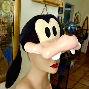 May include: A plush Goofy character hat featuring a black fuzzy fabric top with long floppy ears, a moulded plastic face with a large snout, a black nose, and two prominent white teeth. The hat is designed to be worn over the head as a costume accessory.