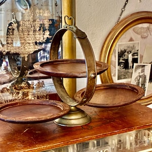 Peut inclure: Présentoir à trois niveaux de style vintage avec plateaux en bois et cadre en métal de couleur laiton. Le présentoir a une poignée et une base décorative. Les plateaux sont ronds et ont une finition marron foncé. Le présentoir est exposé sur une surface en bois.