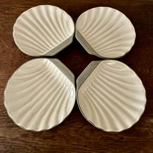 May include: A set of four off-white ceramic plates shaped like scallop shells. Each plate features a series of raised, radiating ridges that mimic the natural texture of a seashell, with a smooth, glossy finish and a scalloped edge design.