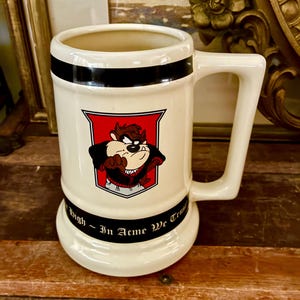 May include: Cream-colored ceramic beer stein featuring a cartoon Tasmanian Devil character in a red and black crest. The stein has a black band near the top and a curved handle. The text "High ~ In Acme We Trust" is printed on the base.