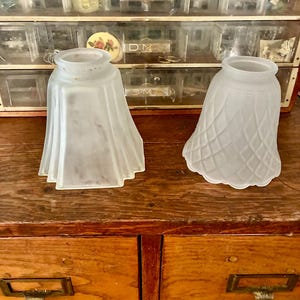 Puede incluir: Dos pantallas de lámpara de vidrio esmerilado. Una pantalla tiene un diseño plisado y la otra un patrón de diamantes. Las pantallas están sobre una superficie de madera. El fondo incluye un armario de almacenamiento vintage con pequeños cajones.
