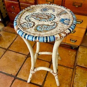 May include: A small, round, white wicker table with a painted top featuring blue and brown paisley designs. The table has four curved legs and a blue and white striped border. The table is on a tiled floor.