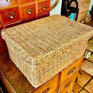 May include: A rectangular storage basket crafted from light brown woven wicker. The basket features a flat, hinged lid that covers the entire top opening, with a tight, uniform weave pattern throughout the sides and lid for a sturdy construction.
