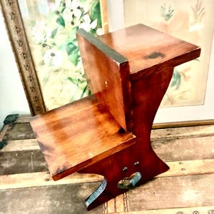 Puede incluir: Un taburete de madera vintage con un rico acabado marrón rojizo. El taburete presenta un diseño de dos niveles con un asiento pequeño y un escalón inferior. Las patas tienen una forma curva única y el taburete parece estar bien elaborado.