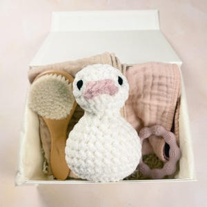 May include: A white crocheted duck toy with black eyes and a pink beak, a wooden hairbrush with soft bristles, a dusty pink muslin cloth, and a mauve silicone teething ring with star and swirl textures, all arranged inside a white gift box.