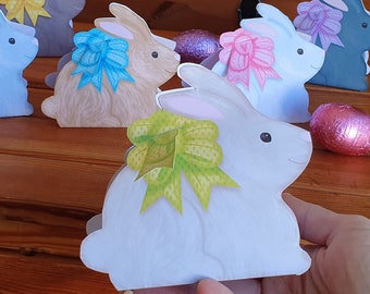May include: Five Easter bunny treat boxes made of paper. The boxes are shaped like bunnies and have different colored bows. The boxes are on a wooden surface. The text "Easter Bunny Treat Box + editable tags" is at the top of the image.
