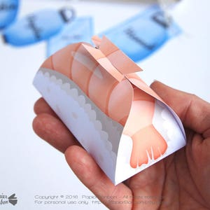 May include: A small, folded paper box shaped like a sushi roll with a shrimp on top. The box is decorated with a white and gray pattern. The text "Yummy Sushi Gift Box & Editable tags" is at the top of the image.