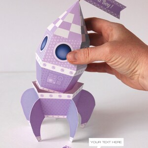 May include: Purple and white paper rocket with a checkered pattern and a flag that says "It's a Boy!"  The rocket has a window and a small panel with a design.  The rocket is sitting on a white surface.  There is a separate flag that says "Welcome Baby Boy!"