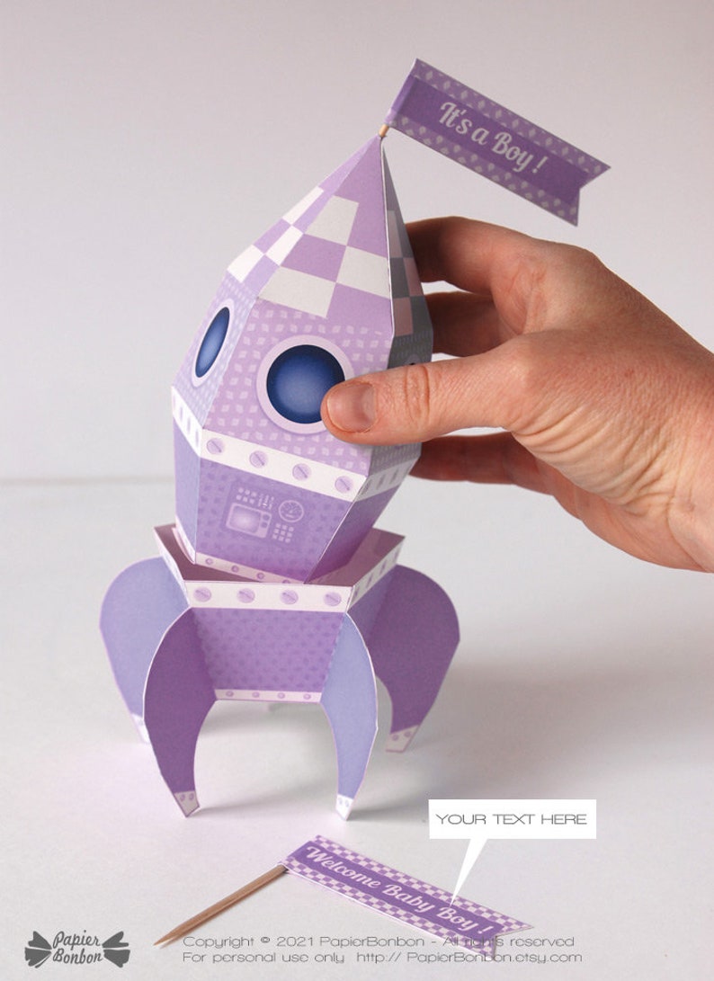 May include: Purple and white paper rocket with a checkered pattern and a flag that says "It's a Boy!"  The rocket has a window and a small panel with a design.  The rocket is sitting on a white surface.  There is a separate flag that says "Welcome Baby Boy!"