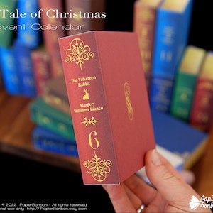 Puede incluir: Una caja roja con forma de libro con letras doradas y un n&uacute;mero 6 dorado. El t&iacute;tulo del libro es "The Velveteen Rabbit" de Margery Williams Bianco. La caja es parte de un calendario de adviento.