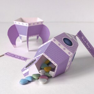 May include: Purple rocket-shaped party favor boxes with white polka dots and a blue window. The boxes are filled with pastel-colored candy hearts and have a flag that says "It's a Boy!"