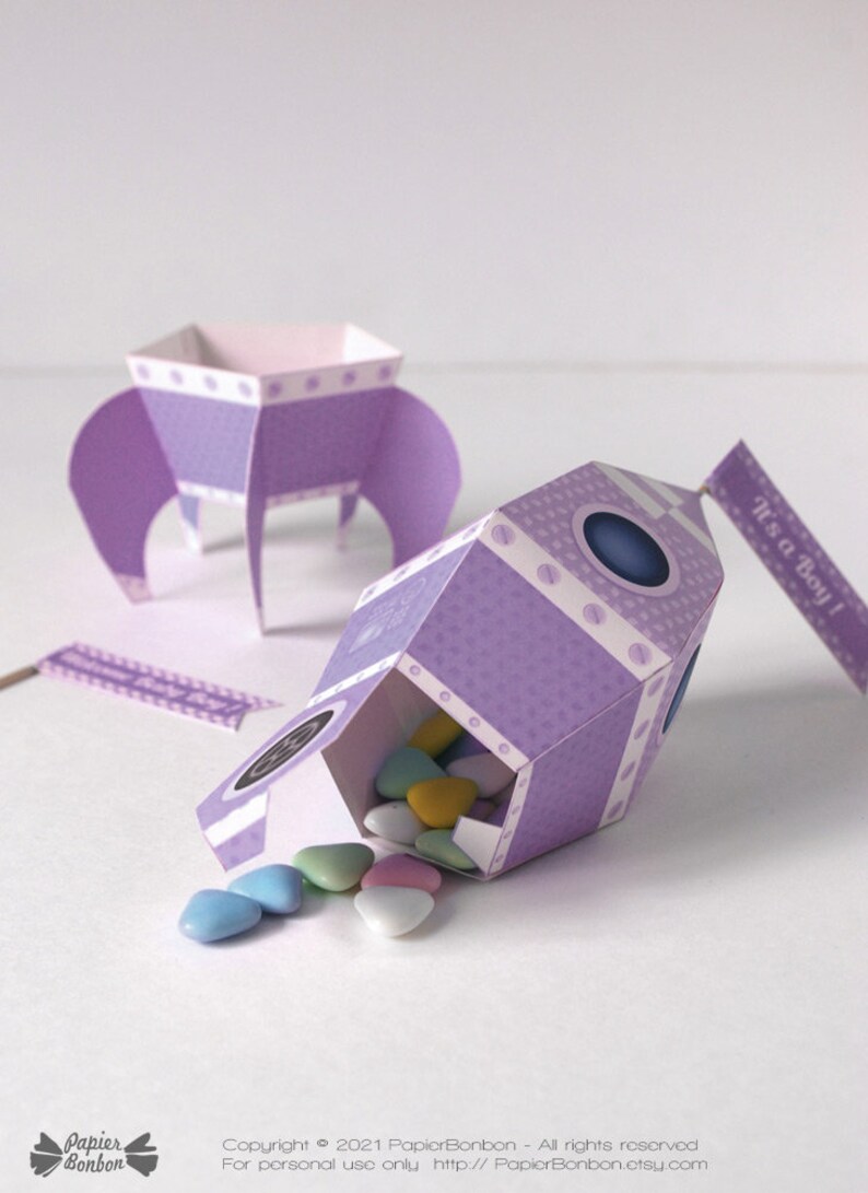 May include: Purple rocket-shaped party favor boxes with white polka dots and a blue window. The boxes are filled with pastel-colored candy hearts and have a flag that says "It's a Boy!"