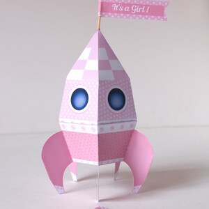 May include: A pink paper rocket with a white and pink checkered pattern, blue eyes, and a pink flag that says "It's a Girl!"