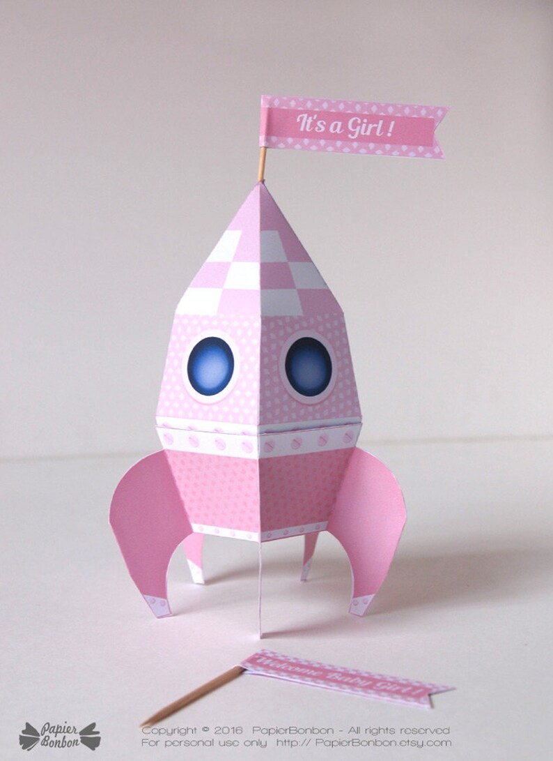 May include: A pink paper rocket with a white and pink checkered pattern, blue eyes, and a pink flag that says "It's a Girl!"