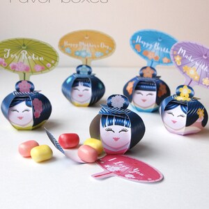 May include: Five colorful Kokeshi paper doll favor boxes with different messages on the top. The boxes are open and filled with small candies. The boxes are decorated with flowers and have a blue and white striped pattern.