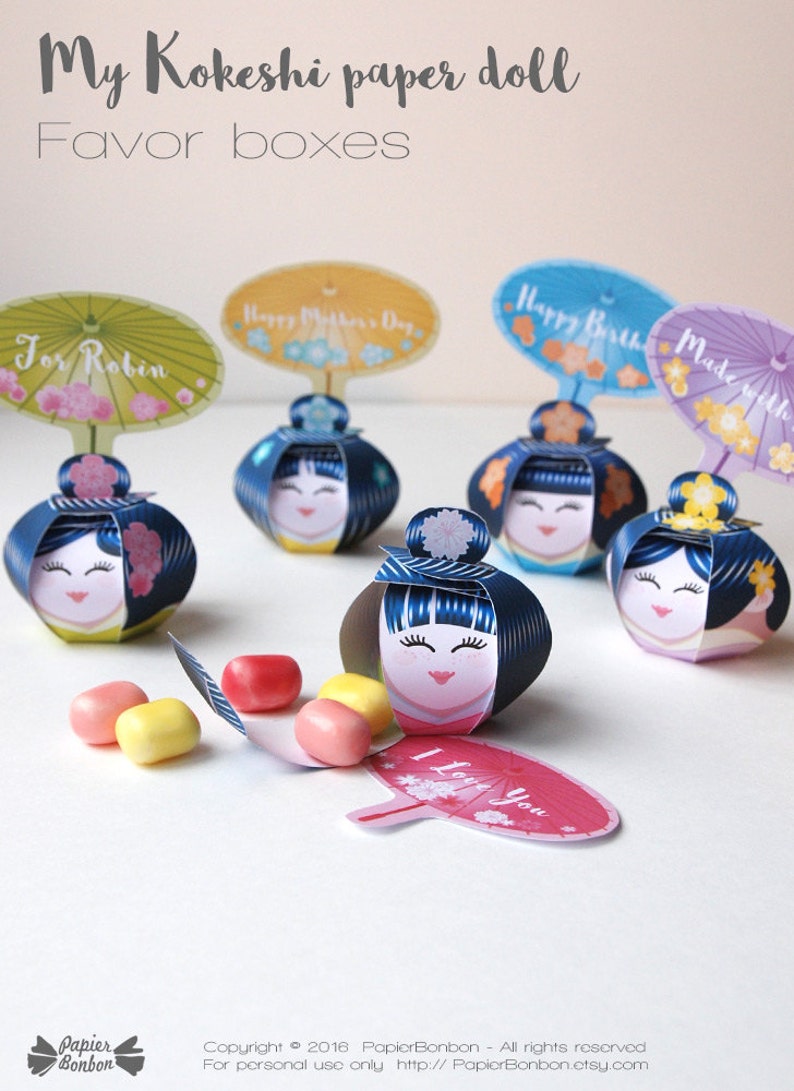 May include: Five colorful Kokeshi paper doll favor boxes with different messages on the top. The boxes are open and filled with small candies. The boxes are decorated with flowers and have a blue and white striped pattern.