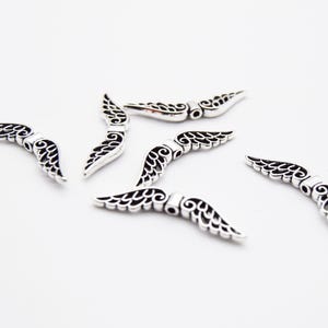 May include: Silver-coloured wing-shaped charms with detailed openwork patterns. The charms are scattered on a white surface, highlighting their delicate design. Ideal for jewellery making or craft projects.