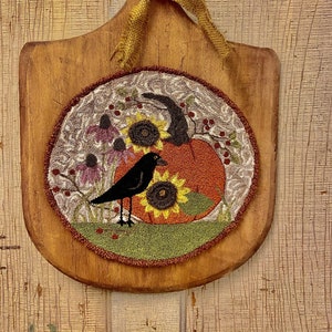 May include: A wooden pizza peel with a round, embroidered design featuring a black bird, a pumpkin, and sunflowers. The design is framed with a brown border and a green outer edge.