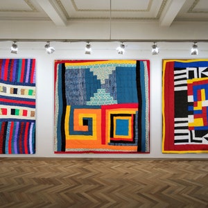 Könnte beinhalten: Drei große, farbenfrohe Quilts hängen in einer Galerie an einer weißen Wand. Die Quilts weisen geometrische und abstrakte Designs in verschiedenen Farben auf, darunter Rot, Blau, Gelb und Schwarz. Die Quilts werden von Schienenbeleuchtung beleuchtet, und der Boden ist aus Holz.