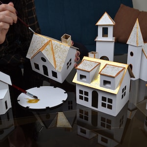 May include: A collection of miniature white houses with gold accents, some are being painted with a brush and gold paint. The houses are on a black surface.