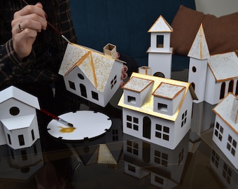 May include: A collection of miniature white houses with gold accents, some are being painted with a brush and gold paint. The houses are on a black surface.